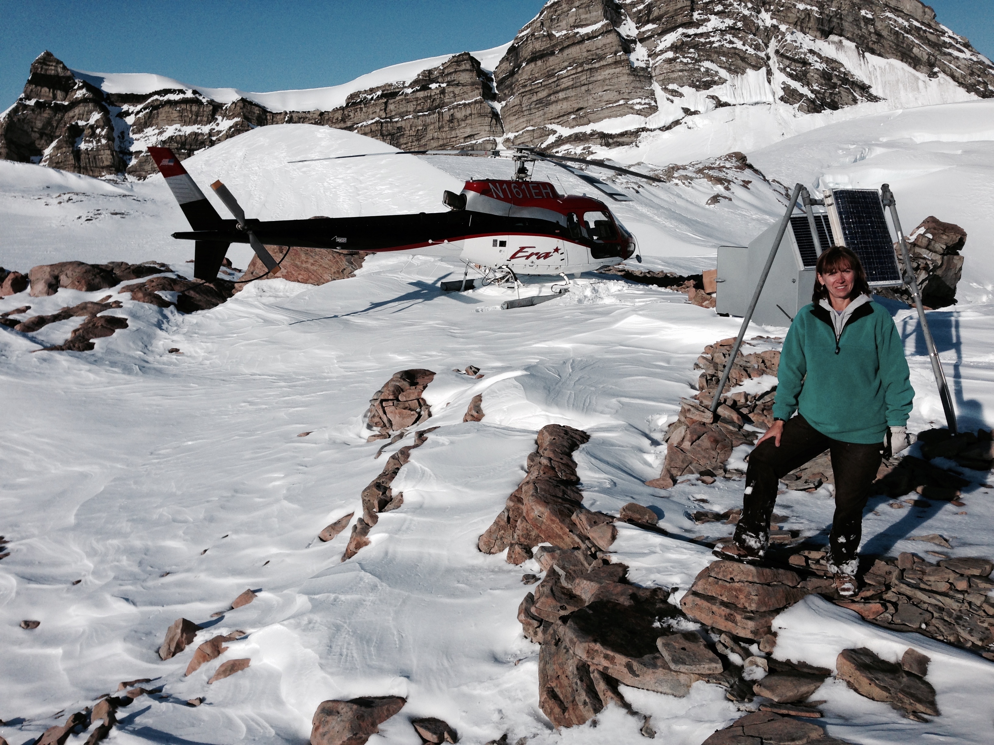 Natalia Ruppert stands on snowy mountain in front of helicopter and seismic site. 