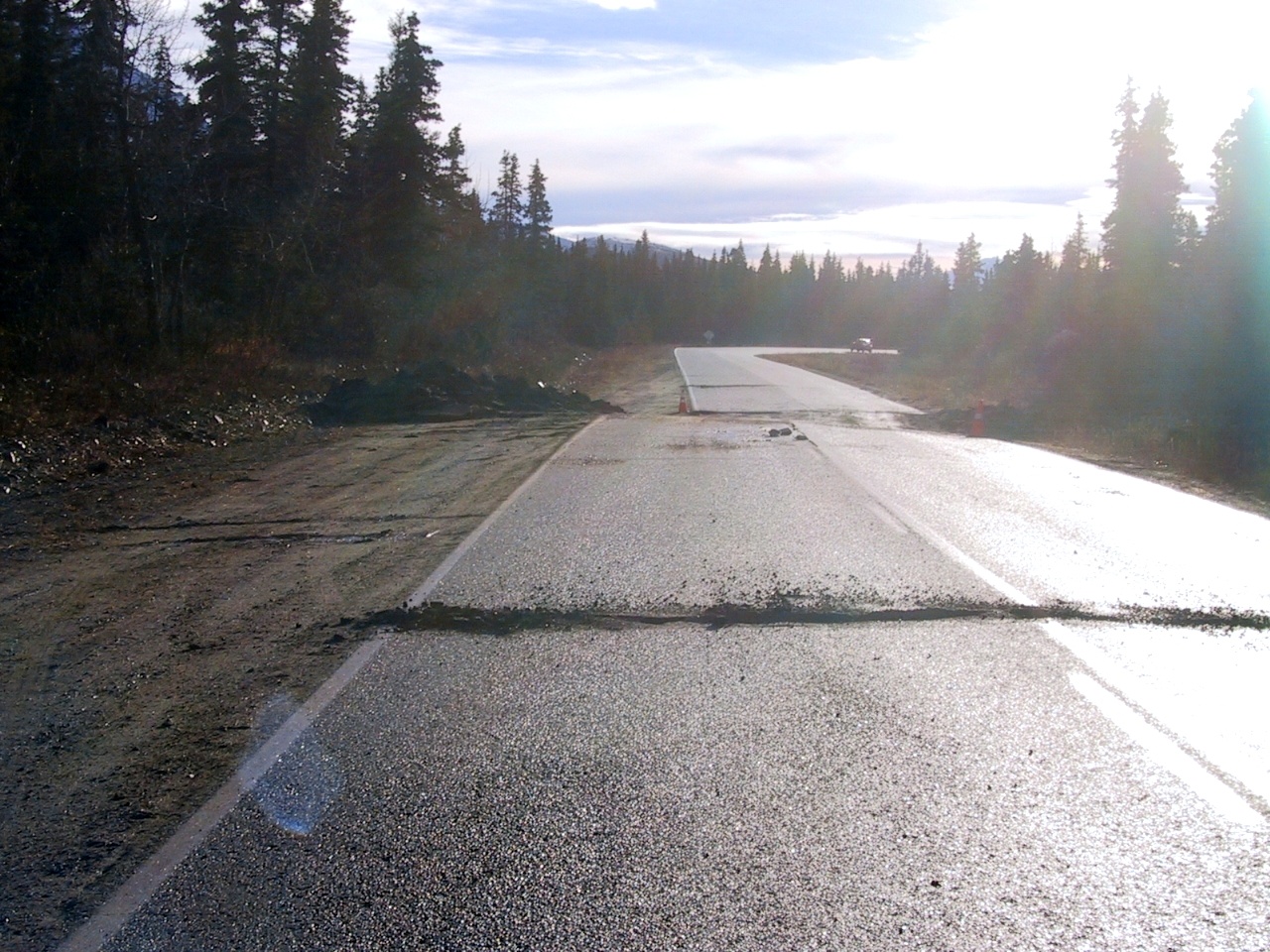 A 2-lane highway extends from the photo foreground to the background, with one car in the distance and flanked by thick spruce forest. In the mid-ground, earthquake damage crosses the width of the road, with one part of the road offset by several feet from the section on the other side.