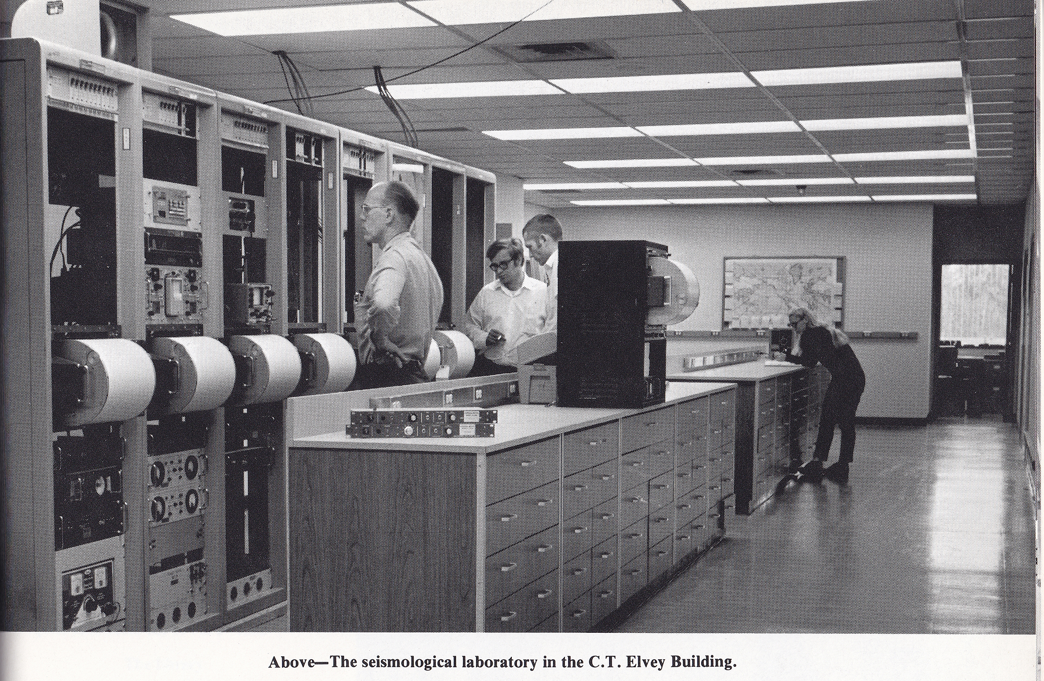 Black and white photo of tall row of seismometers with paper drums, with people looking at machines.