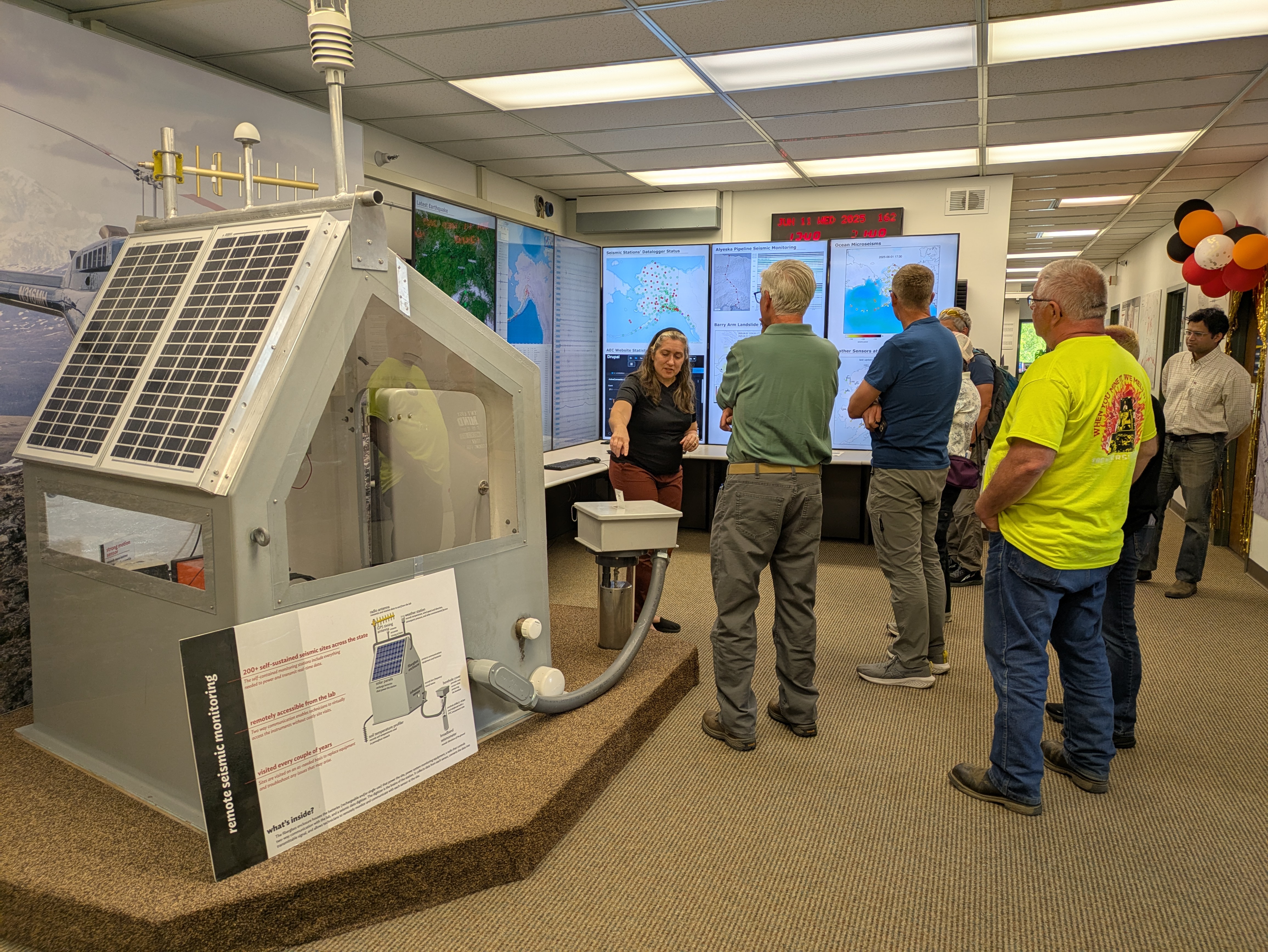 Woman pointing at seismic station hut, group of people standing in front of her.