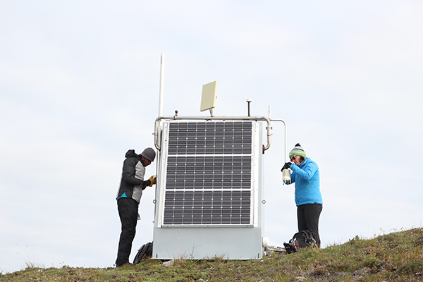 Two people on either side of a station hut installing a meteorological sensor. 