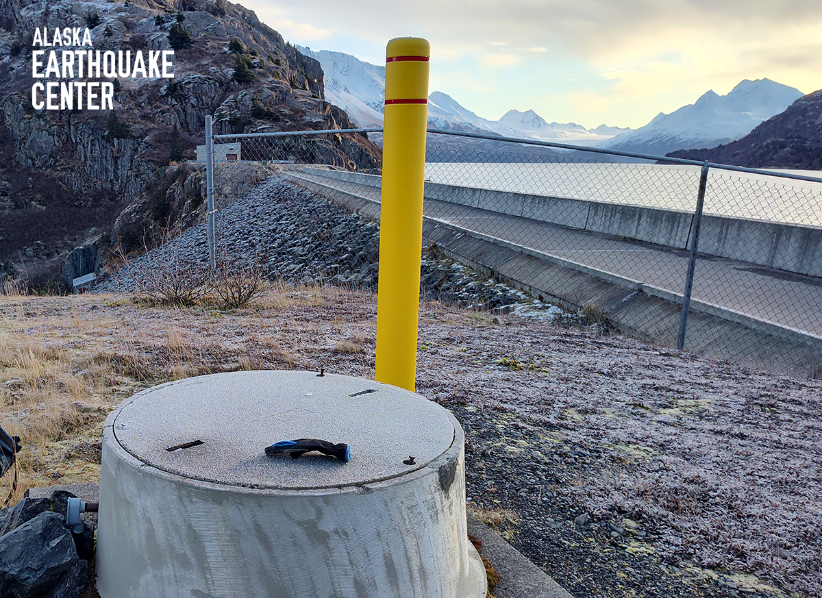 Concrete cylinder in foreground is the vault housing the seismometer. A chain-link fence blocks the top of the dam, which extends to the background.