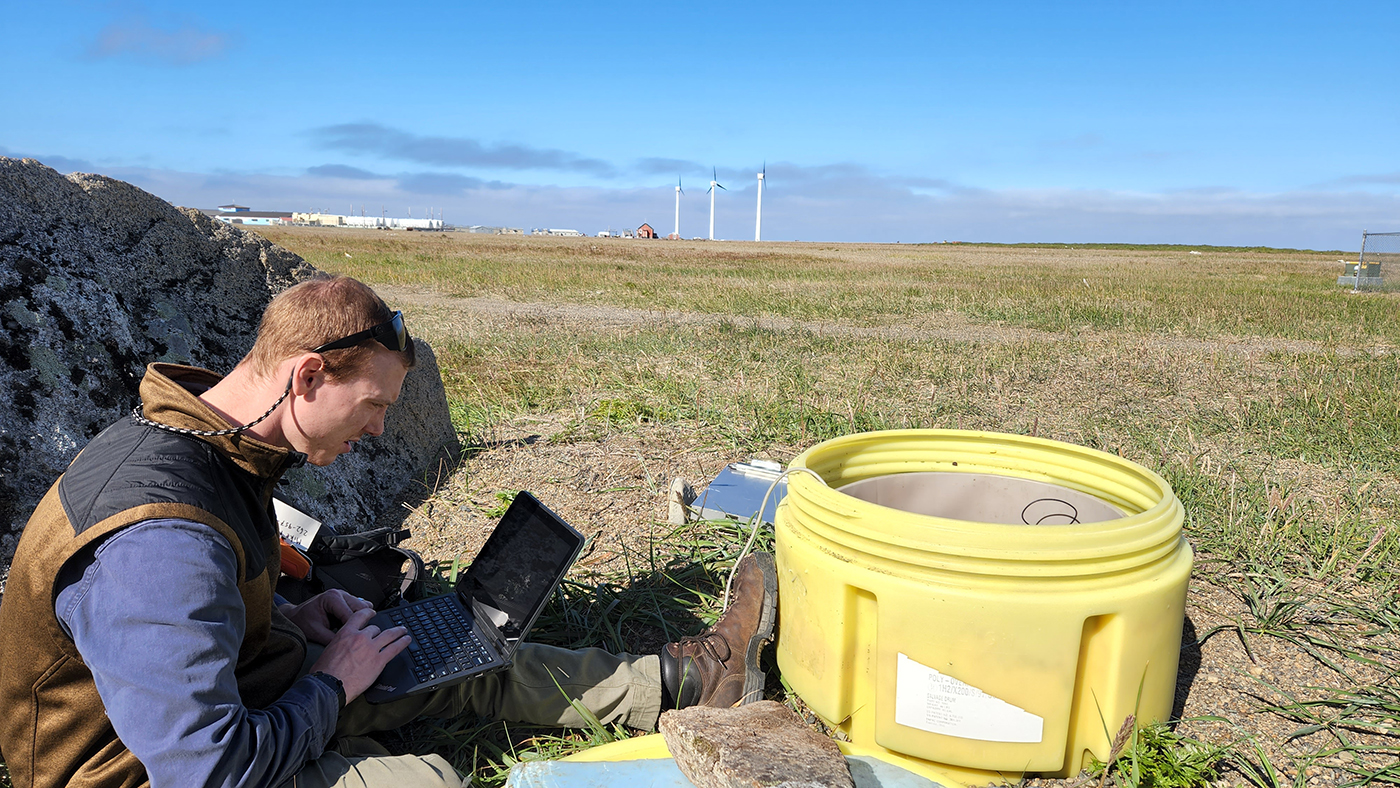 Man sitting on ground next to plastic seismometer vault and working on laptop. 