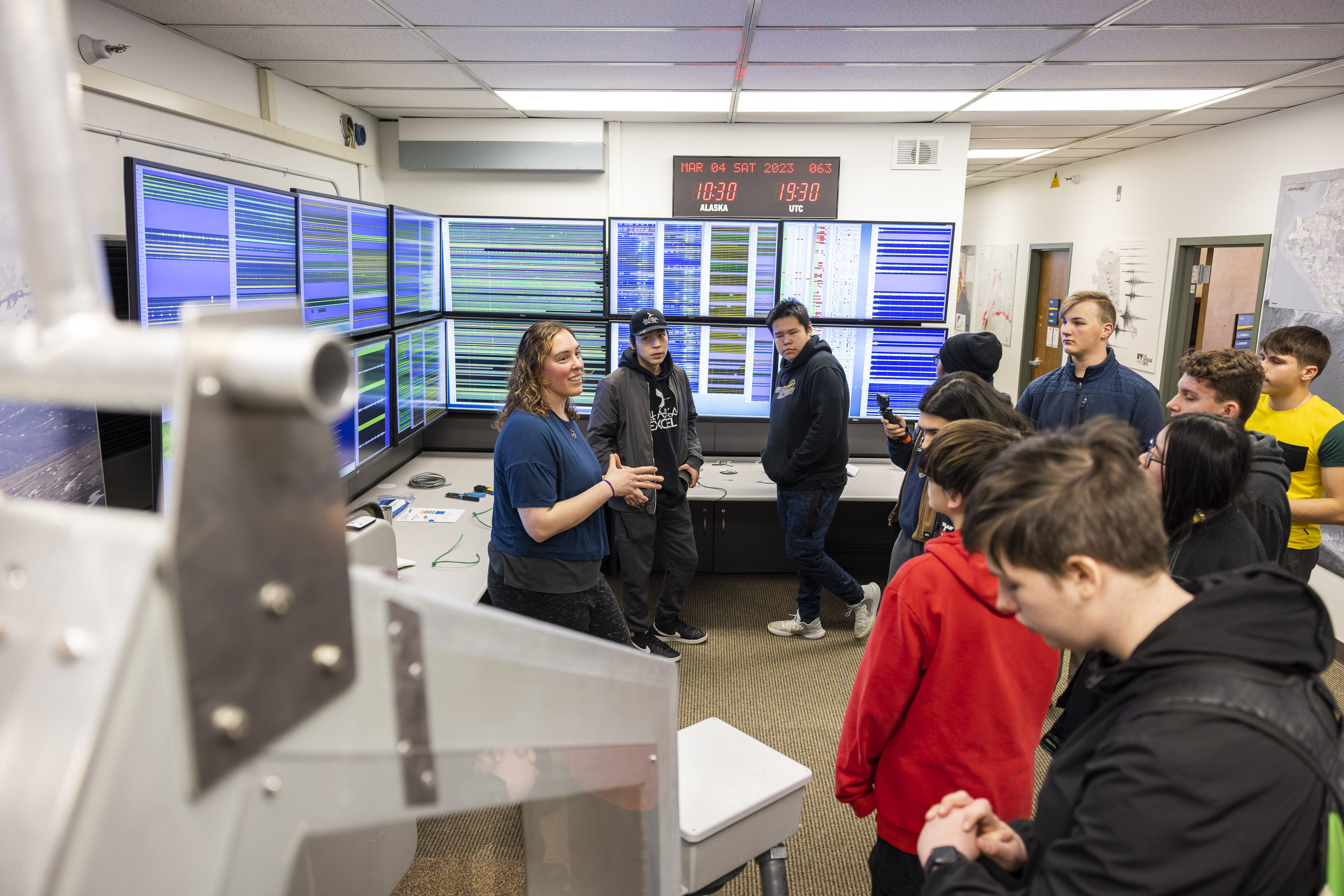 Female staff member standing in front of multiple seismograph monitors, talking to a group of high school students.