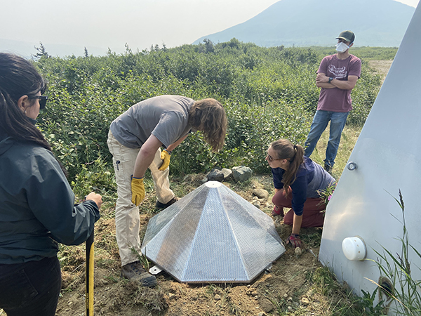 Two people standing watching and two people bent over installing cone-shaped wind noise reduction screen for infrasound instrument.