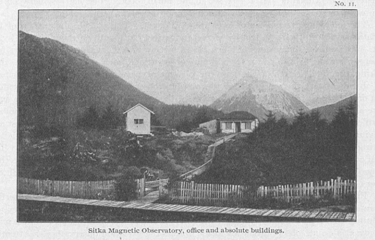 Old black-and-white photo of two white buildings behind a white fence with mountain in the background.
