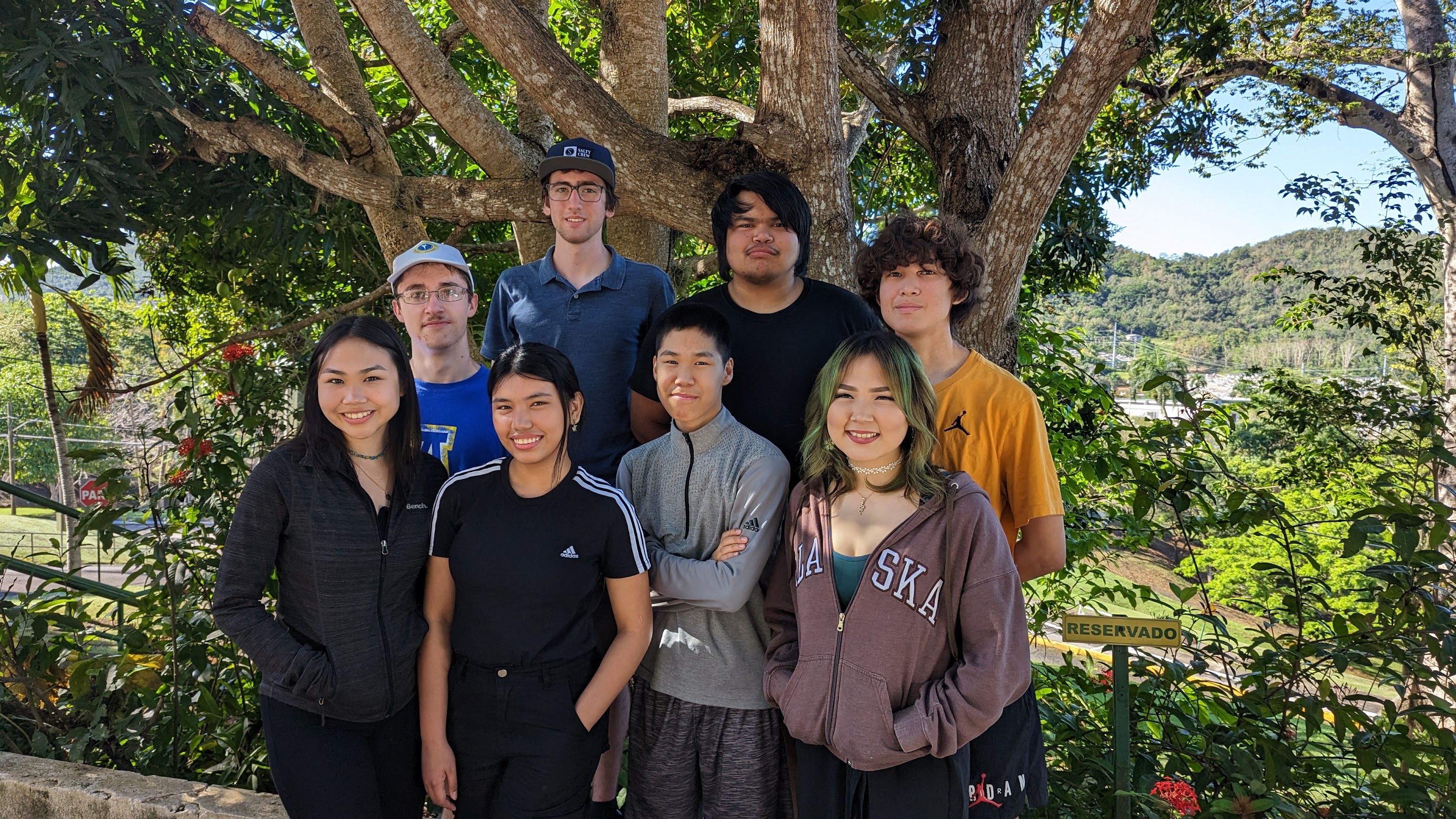 Eight high school students standing in front of tropical foliage and tree. 